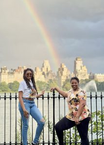 Two women in Central Park smiling with a rainbow in the sky behind them